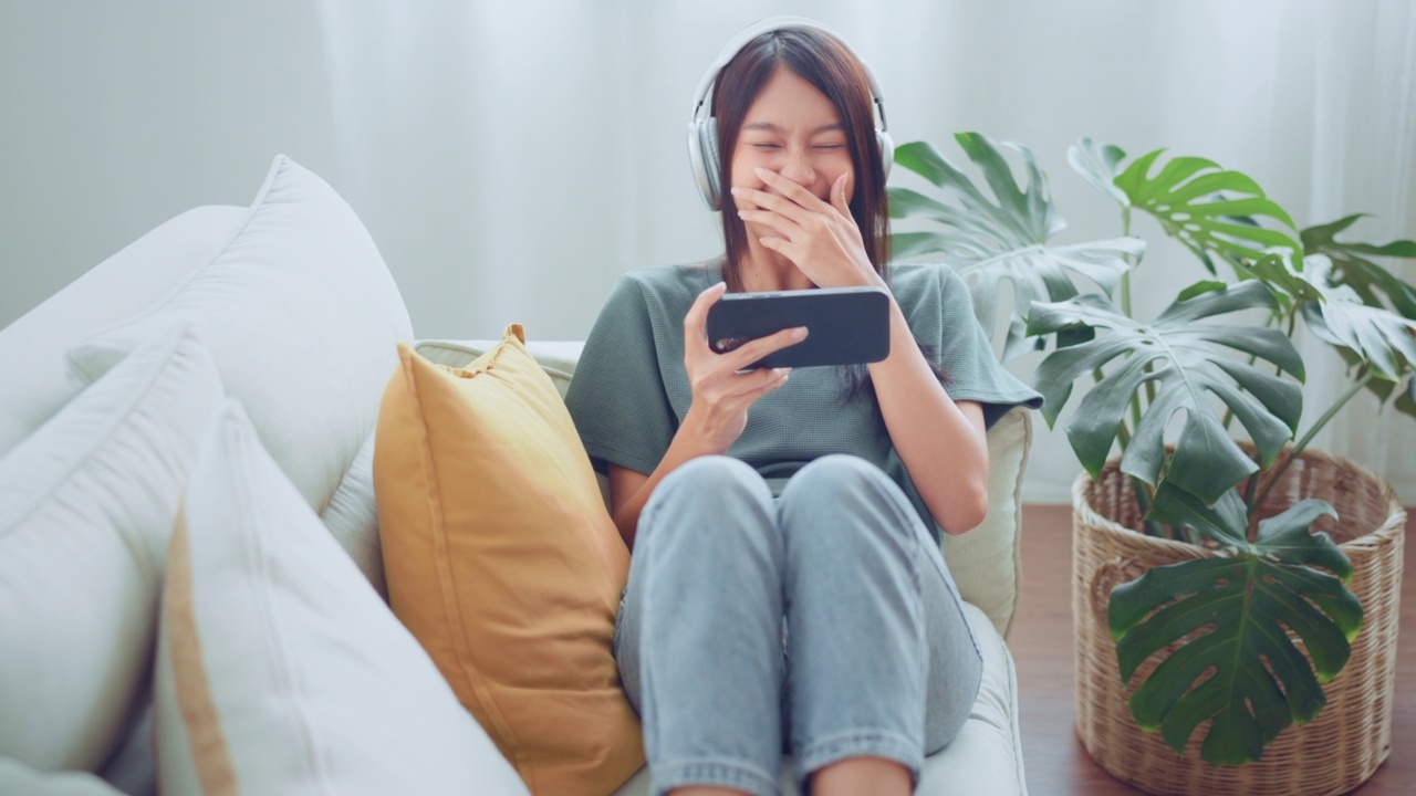 快乐的年轻亚洲女性放松和听音乐和看电影的手机与耳机，而坐在沙发上在家里早上视频素材