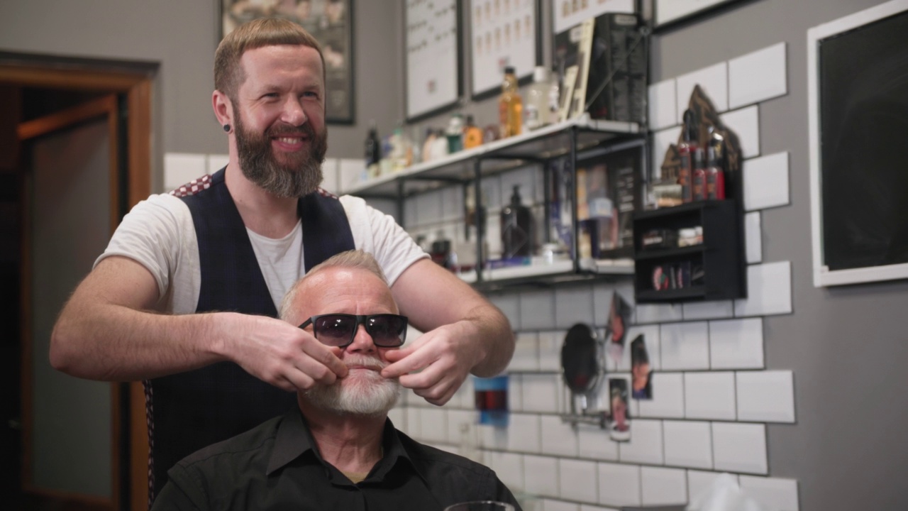 一位戴眼镜的退休老人在理发店里理发和造型，坐在主人的扶手椅上，一位专业的理发师在沙龙里为一个成熟的男人留胡子，对着镜子视频下载