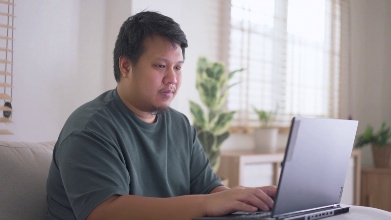 亚洲男人坐在家里的沙发上使用笔记本电脑视频素材