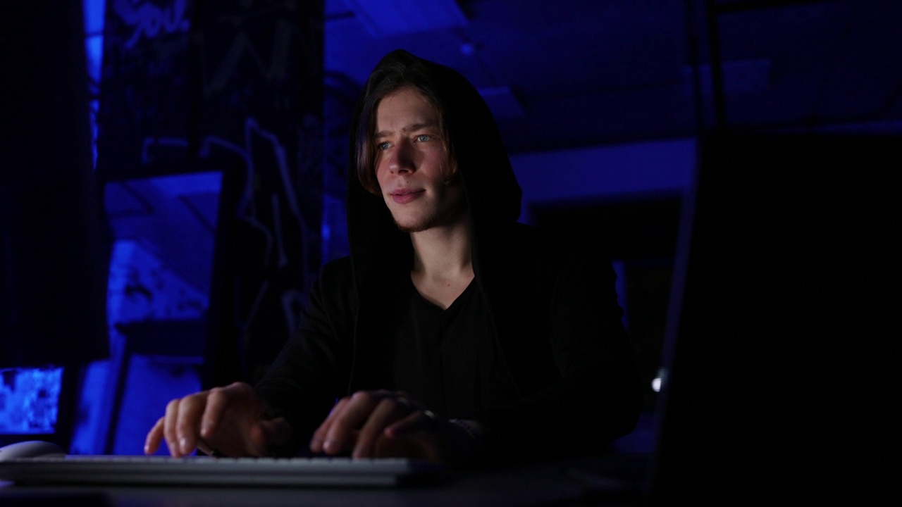 一名隐藏的黑客穿着带兜帽的运动衫，坐在有蓝色霓虹灯的黑暗房间里，正在入侵安全系统。视频素材