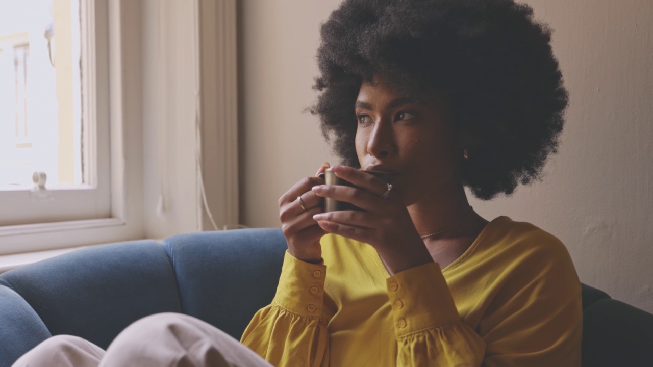 年轻轻松的女人坐在沙发上喝咖啡在休息室在家里。一位扎着爆炸头的满足女性在沙发上放松，早上喝茶。女人享受温暖的饮料视频素材