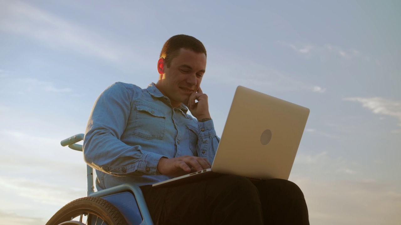 坐在轮椅上的年轻人坐在美丽的天空下，在笔记本电脑前工作。一个残疾人在海滩上用笔记本电脑工作。远程工作. .视频下载