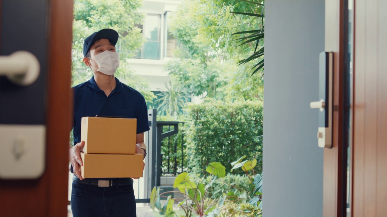 年轻的亚洲快递员戴着面具，穿着蓝色海军制服携带箱子箱子敲门回家网上购物纸箱给女人在门口女士扫描二维码支付在家里的智能手机。视频素材