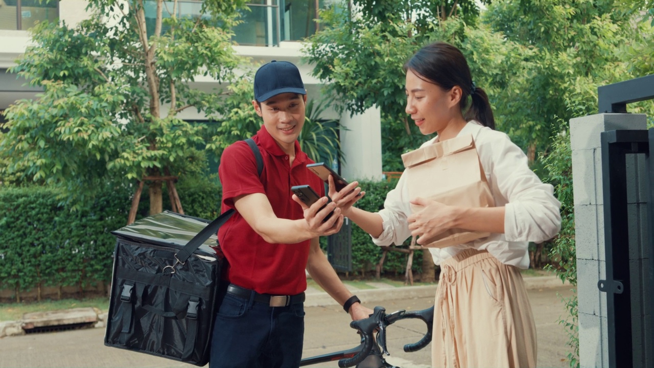 年轻的亚洲快递员骑着自行车，穿着红色制服，携带箱子，敲门回家，网上购物纸袋，女人在门前，女士扫描二维码支付在家里的智能手机。视频下载
