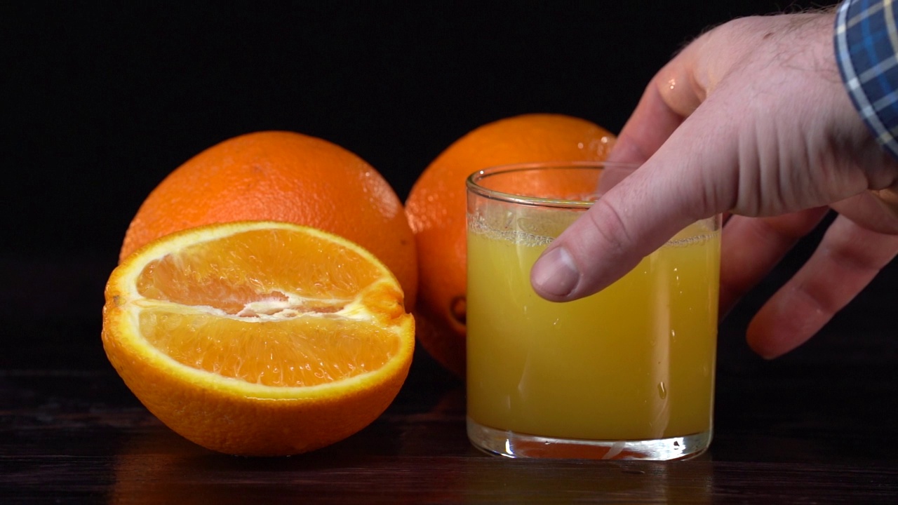 一杯健康的橙汁和橘子。白人手接过杯子，然后把它放在桌子上。餐饮概念。黑色背景。黑暗反映表。慢动作视频下载