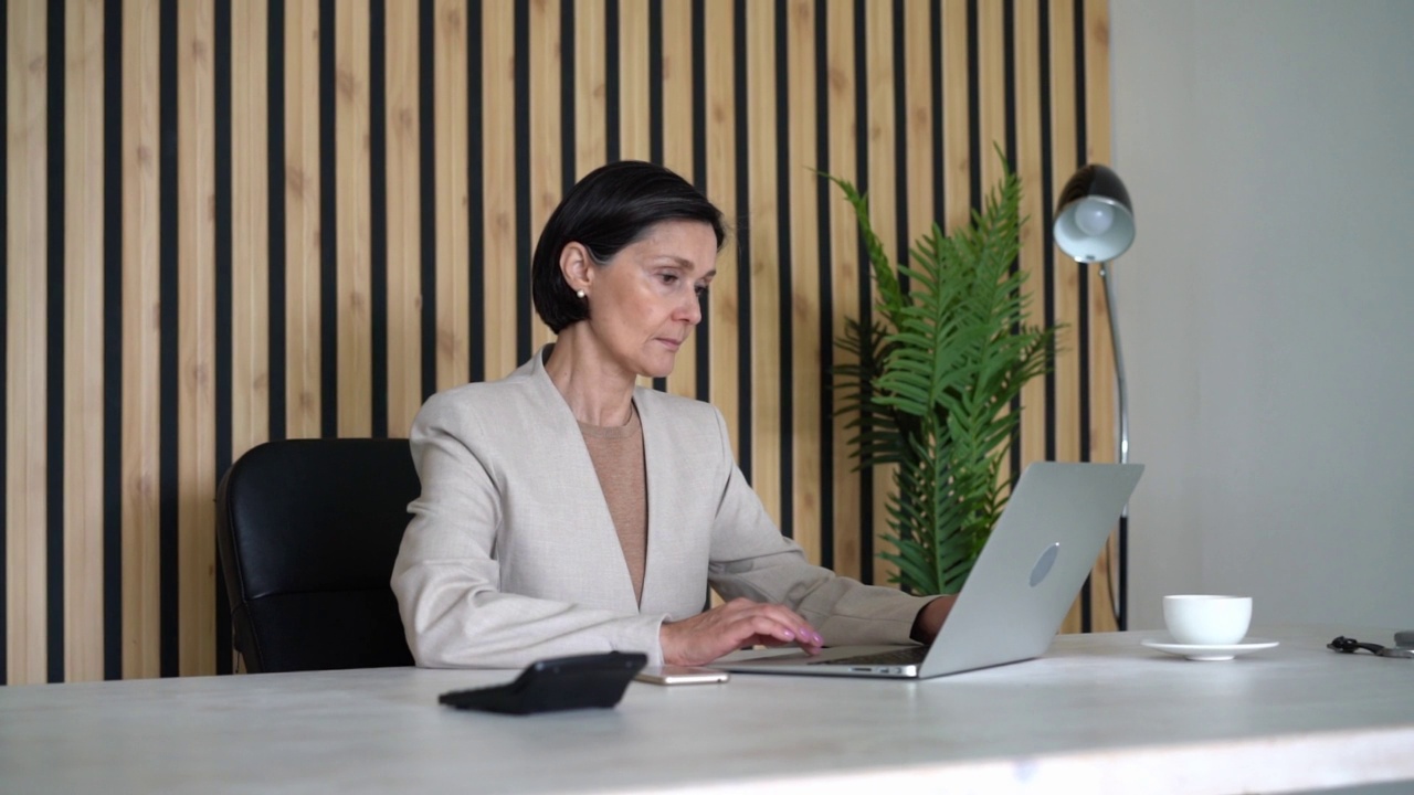 女商人在办公室使用笔记本电脑。高级职员穿着正装，在商务中心办公，坐在办公桌前打字。企业劳动的概念。忙碌的女性在做研究时盯着屏幕。水平、视频视频下载