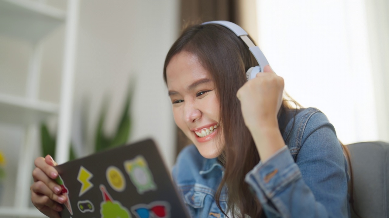 一个戴着白色无线耳机的快乐的年轻女人坐在沙发上，看着她的笔记本电脑庆祝网上的胜利，快乐地捶打着空气。视频素材