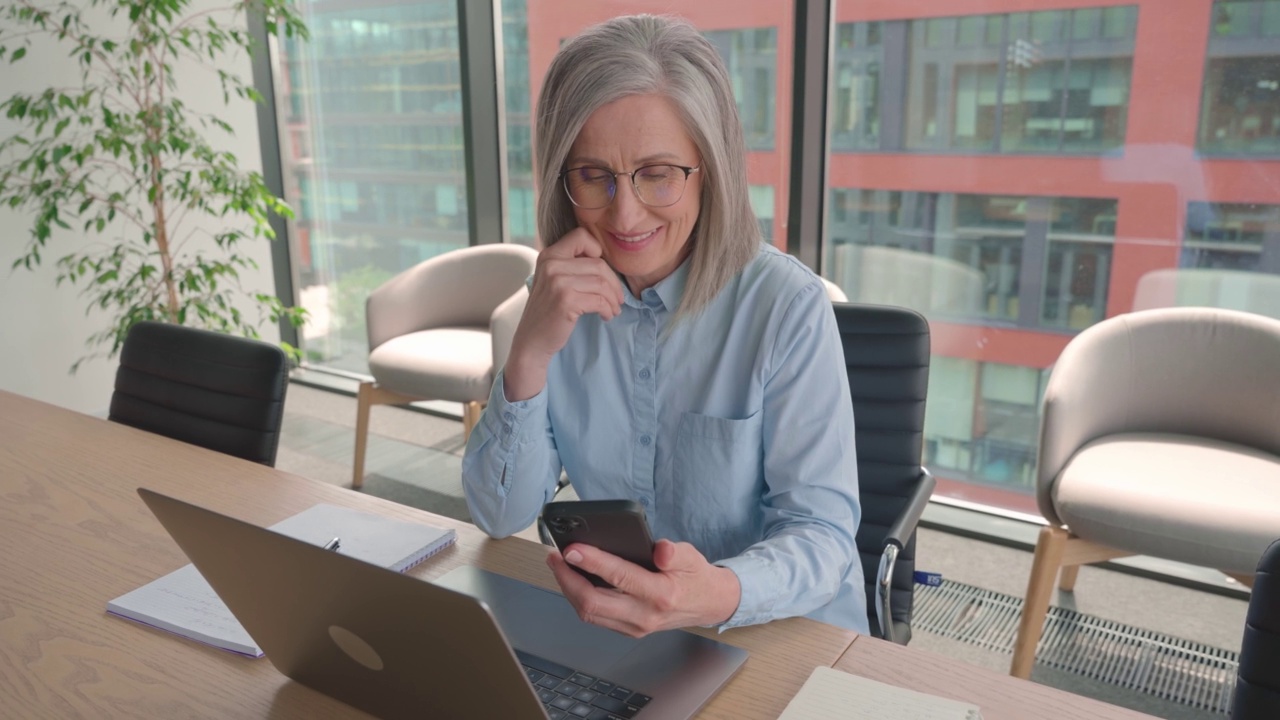 微笑成熟的商业女性拿着智能手机坐在办公室。视频素材