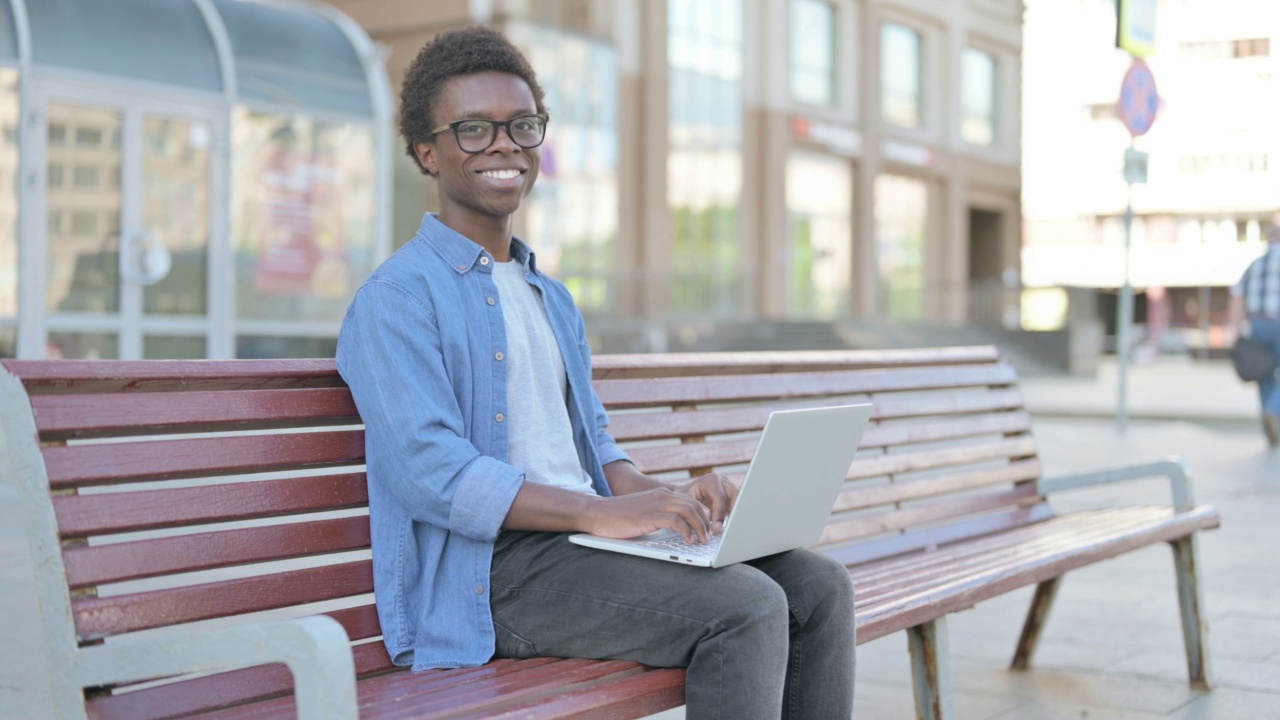 非洲年轻人坐在户外的长凳上，带着笔记本电脑对着相机微笑视频素材