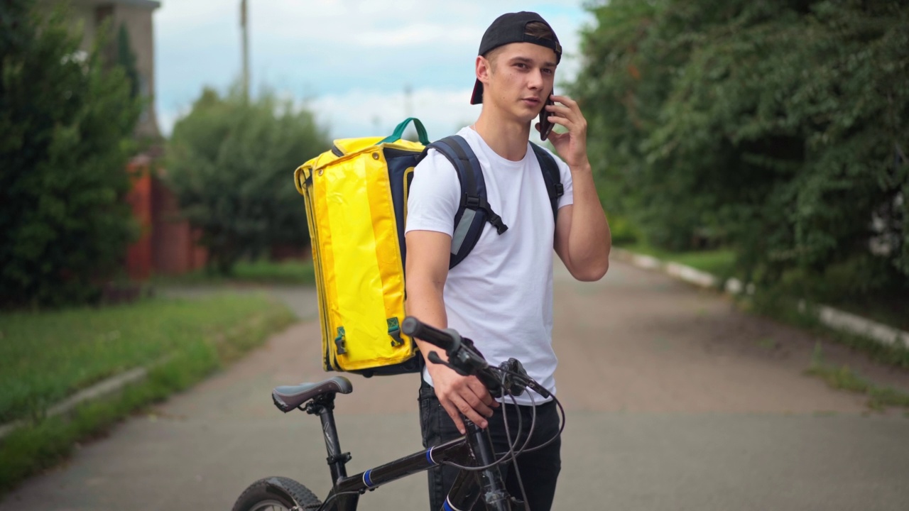 一个白人快递员拿着自行车和黄色的快递袋站在郊区的马路上给客户打电话。年轻英俊的快递员在城里寻找顾客的肖像。视频素材