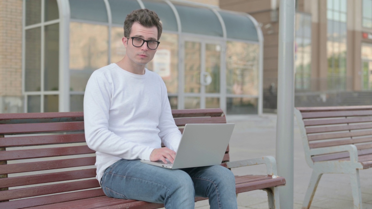 带着笔记本电脑的年轻人坐在户外的长凳上对着镜头微笑视频素材