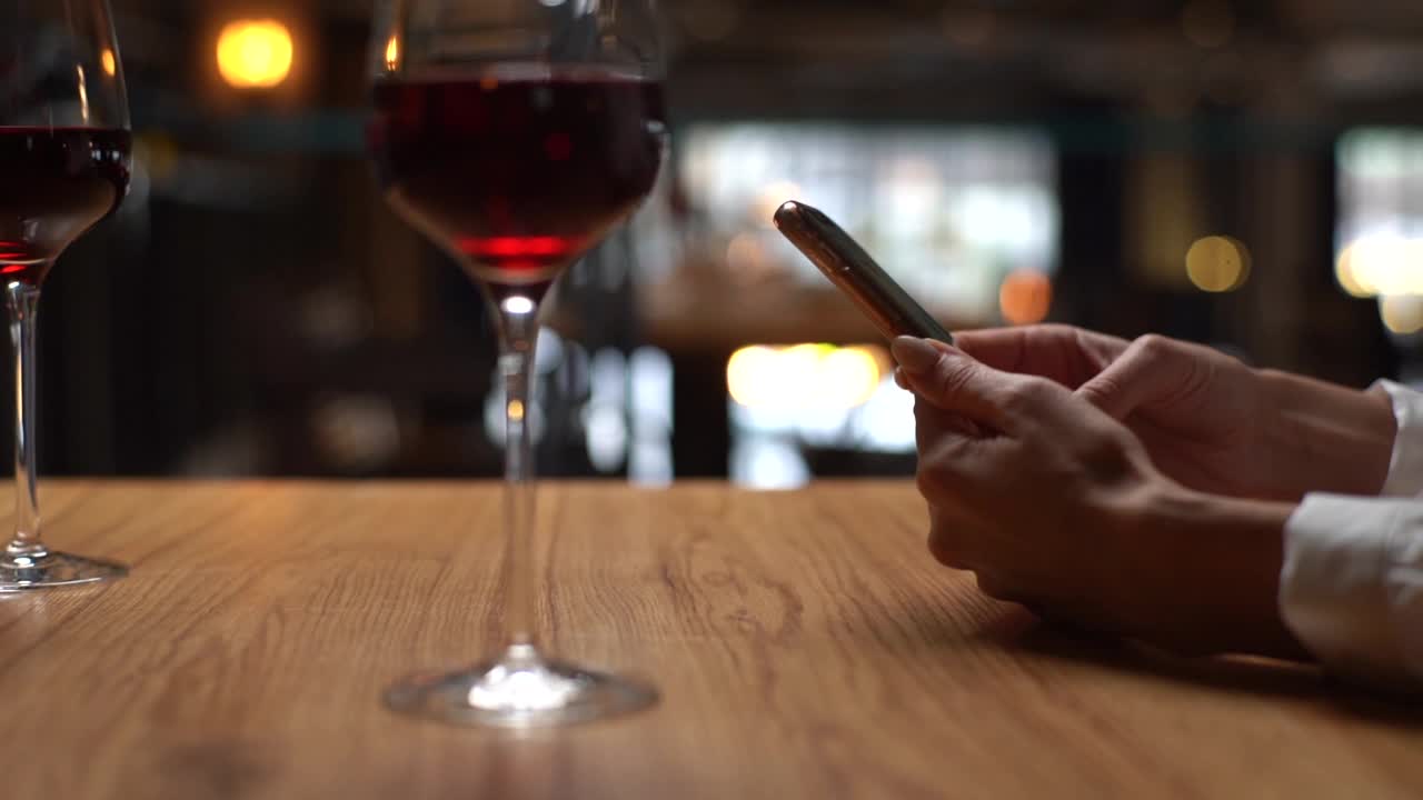 特写，一个陌生的年轻女人坐在桌子旁，拿着几杯酒，在餐厅等男朋友，用智能手机输入信息。视频下载