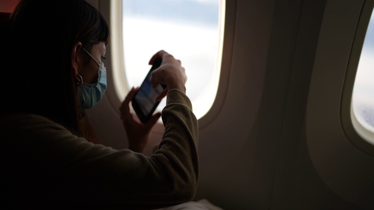 年轻女子戴着口罩坐飞机，透过窗户拍照视频素材