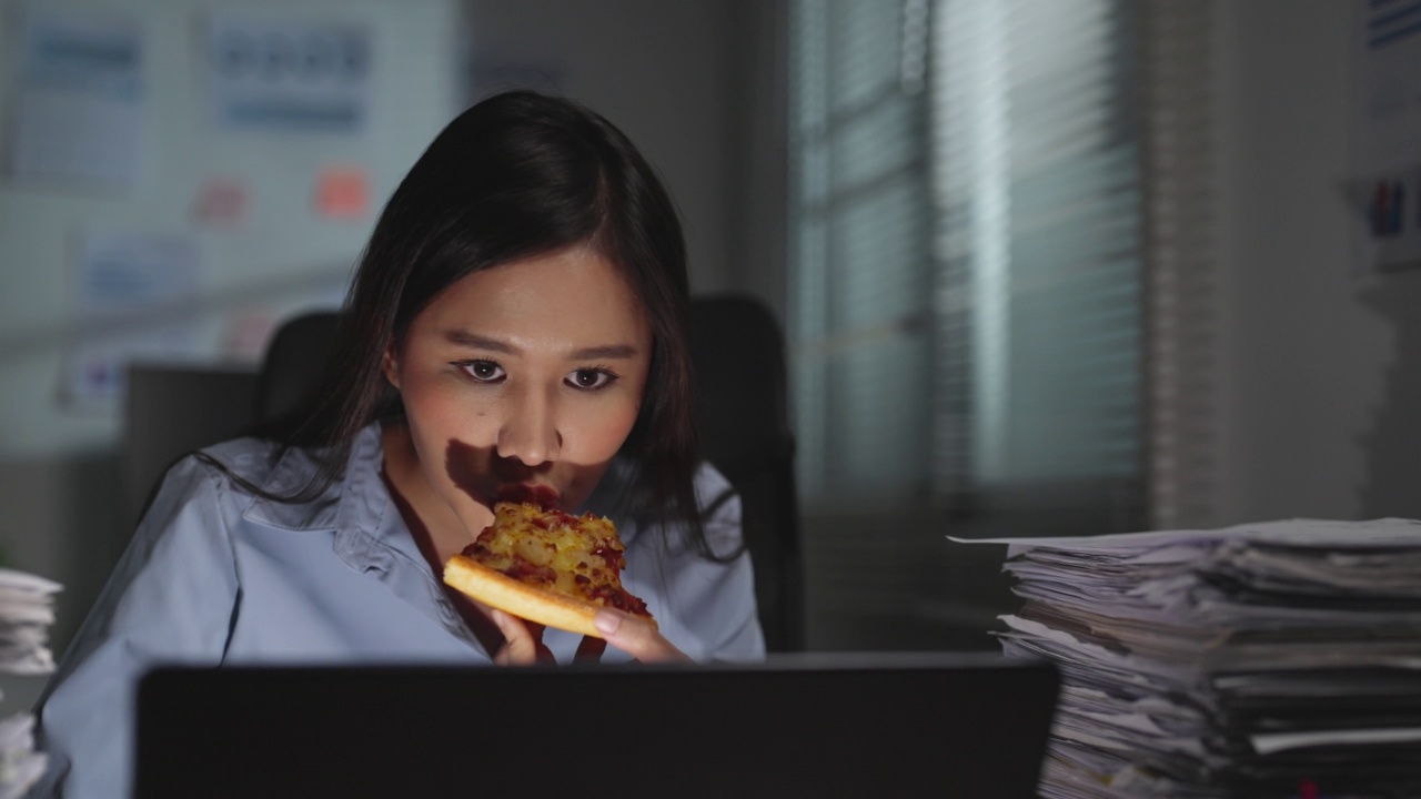 年轻的亚洲女性员工穿着时髦的休闲服装，忙碌地工作着，吃着披萨，在笔记本电脑上完成业务报告，在黑暗的办公室熬夜到深夜。视频下载