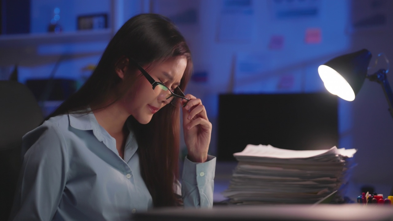 精疲力竭的亚洲商务女性在办公室加班分析报告财务时眼睛疲劳。晚上为清理文件工作到很晚。加班工作到很晚。视频素材