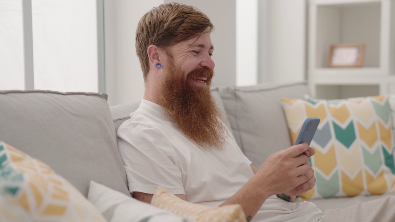 年轻的红发男子坐在家里的沙发上使用智能手机视频素材