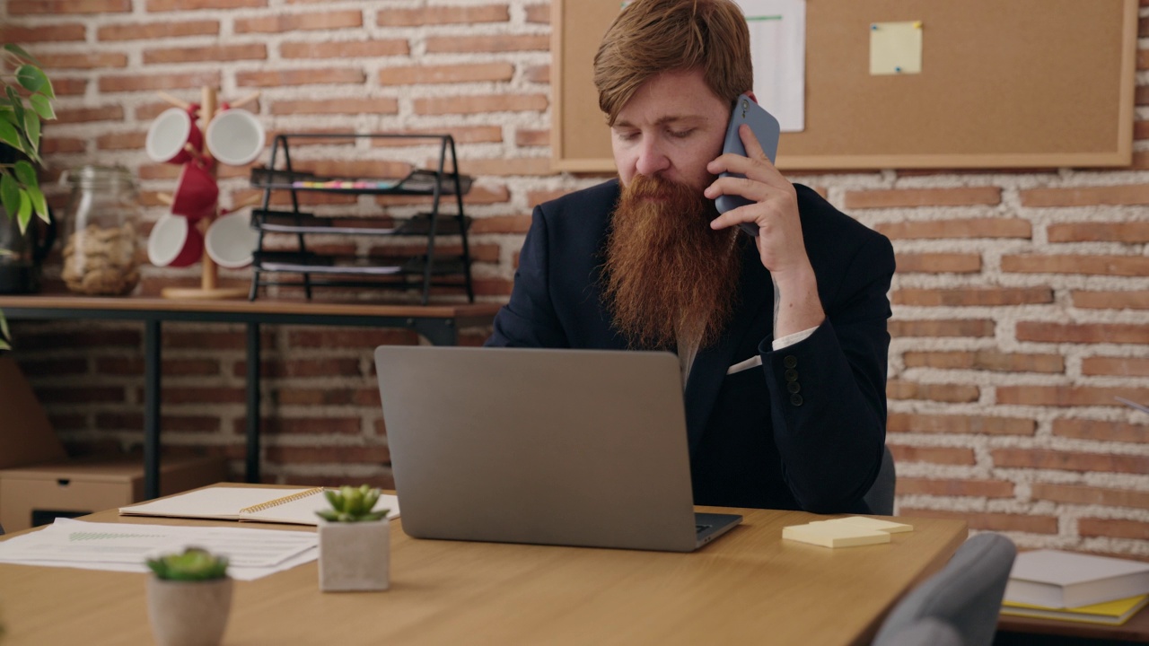 年轻的红发男子商务工作者使用笔记本电脑在办公室用智能手机交谈视频素材
