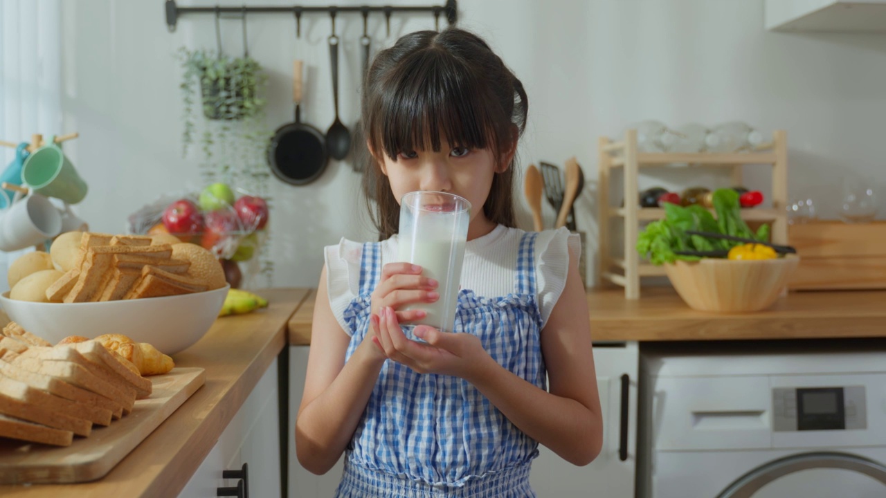亚洲小孩的肖像，在家里的厨房里拿着一杯牛奶和饮料。幼小的学龄前女孩或女儿喝牛奶，弄乱了牛奶的胡子和嘴唇后，在家里的医疗保健。视频素材