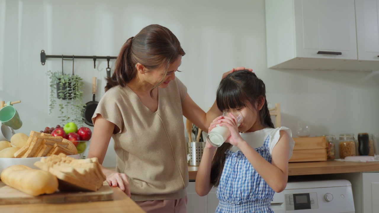 亚洲小可爱的孩子拿着一杯牛奶和喝妈妈。迷人的妈妈教导和支持年轻的女儿照顾她的身体，喝牛奶后醒来在家里的保健。视频素材