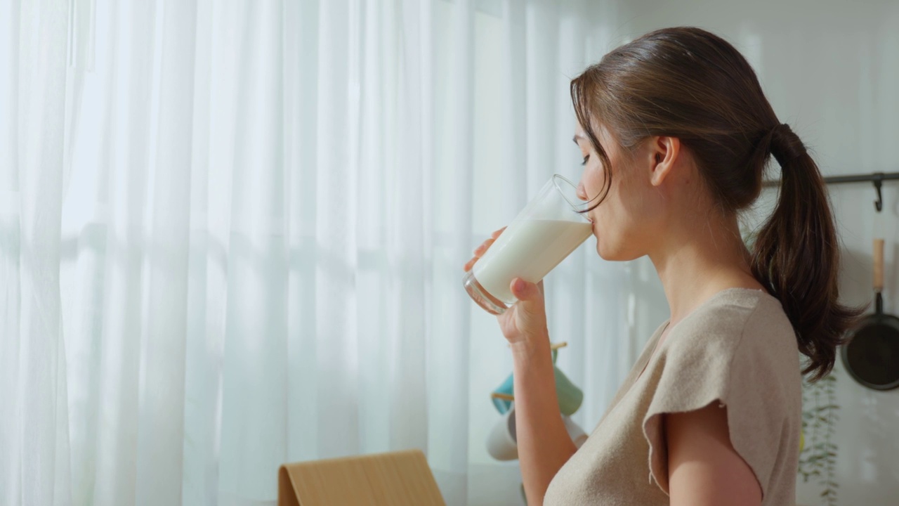 亚洲年轻美丽的女孩喝一杯牛奶在家里的厨房。有吸引力的口渴的女人持有和啜饮一杯牛奶后，早上醒来在家里的健康保健福祉。视频素材