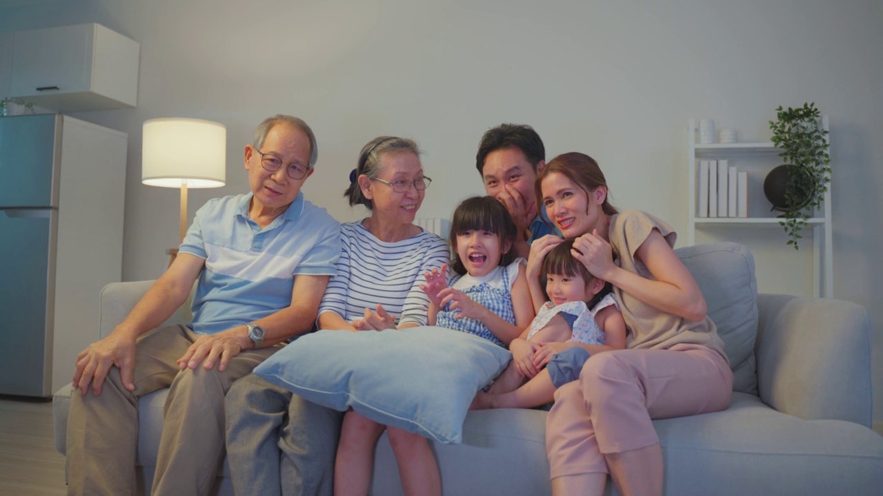 亚洲家庭晚上在家里看电视上的跳楼恐怖电影。年长的祖父母和年轻的夫妇带着小女儿坐在沙发上，看着电视上的恐怖视频，感到兴奋和震惊。视频下载