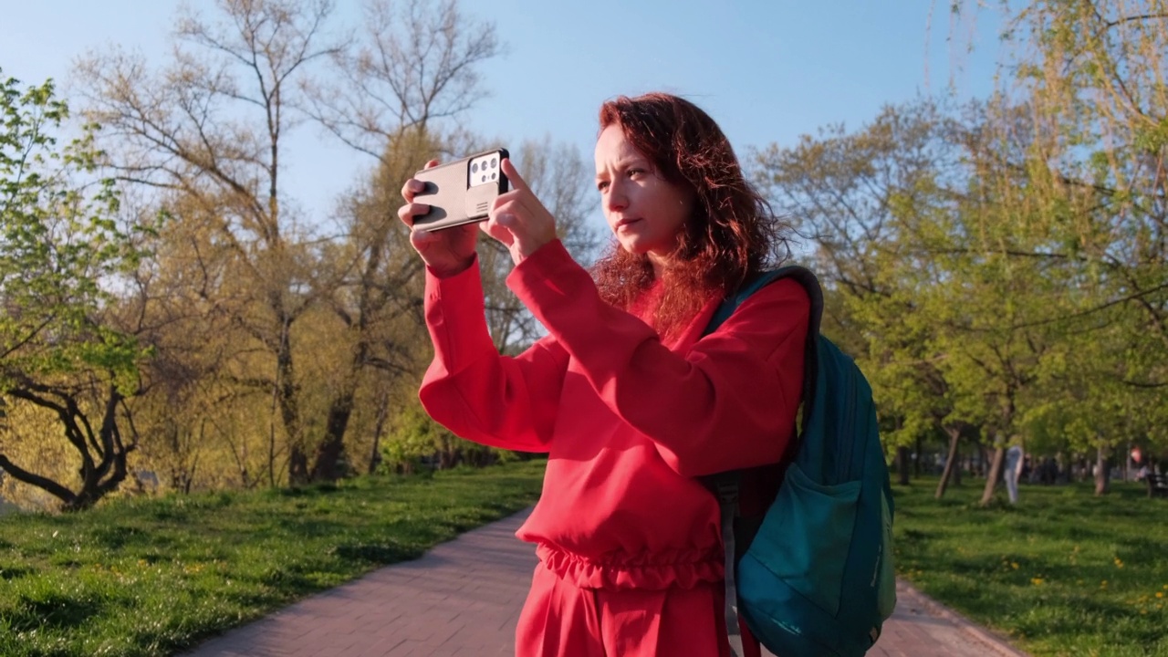 红头发的女人，双手拿着智能手机，正在拍照。公共夏季公园黄金时间视频下载