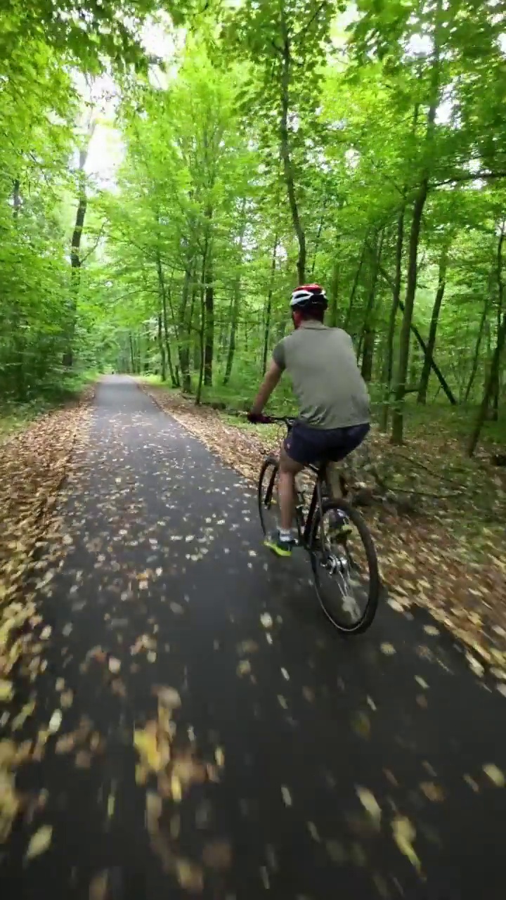一名戴着防护头盔的男子正骑着自行车沿着森林中的柏油路前行。视频素材