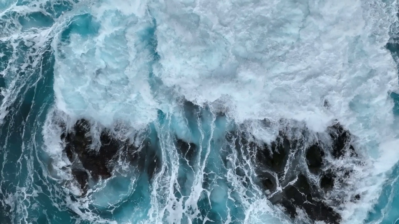 航拍的从上到下拍摄到的海浪拍打在石头上形成白色的泡沫和漩涡。蓝绿色的海水背景，慢动作视频素材