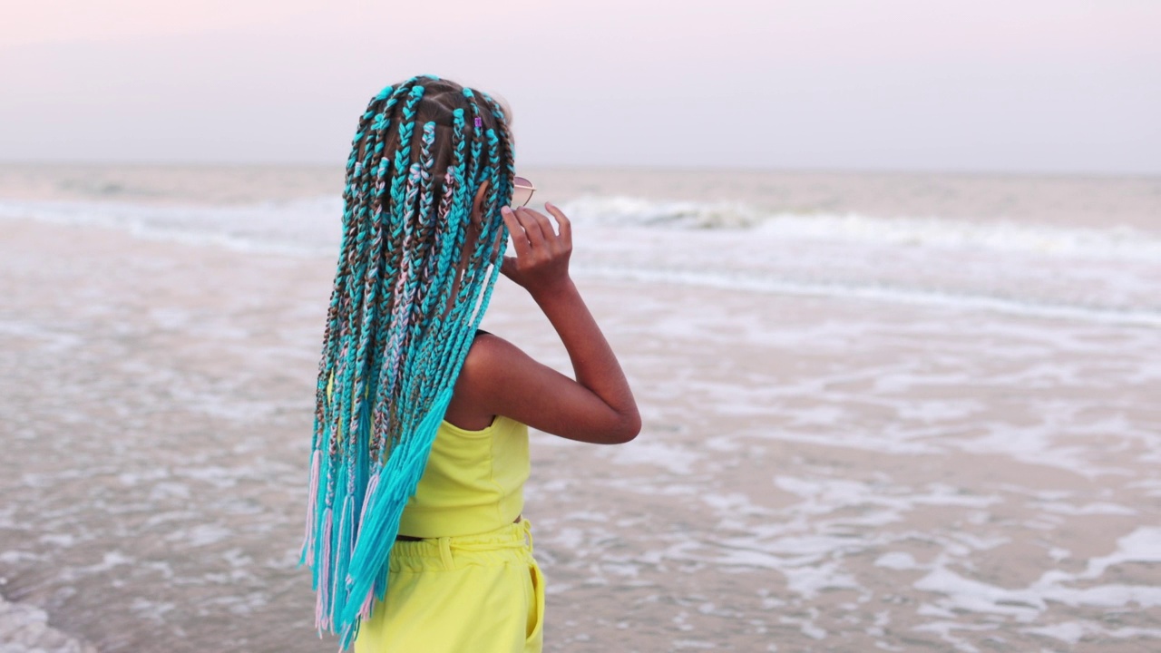 一个扎着非洲麻花辫、穿着夏装的女孩站在海滩上看着大海的地平线视频下载