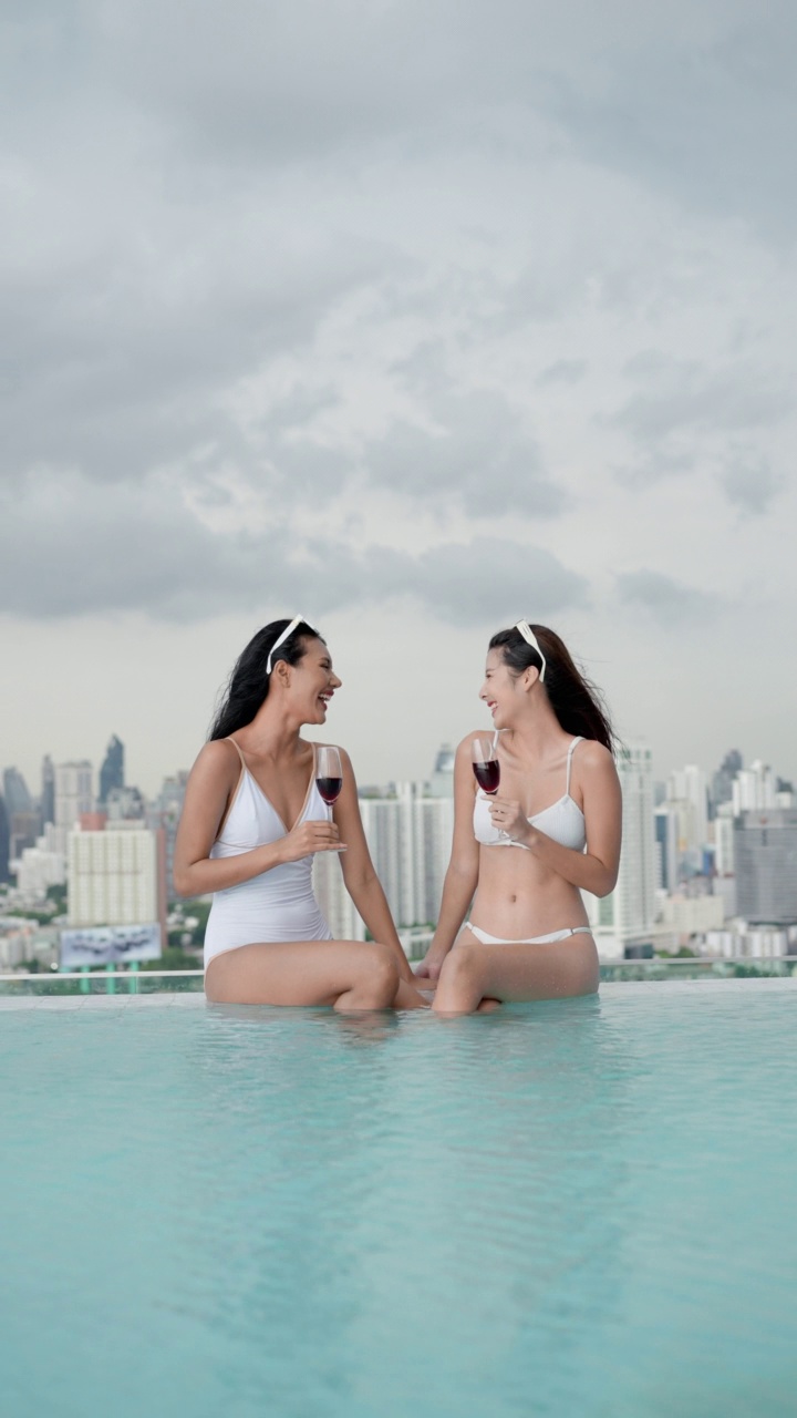两名自信的女性朋友穿着性感的白色比基尼一件两件，手拿红酒坐在泳池边，欣赏着美丽的城市风景。城市背景的性感女人。视频下载
