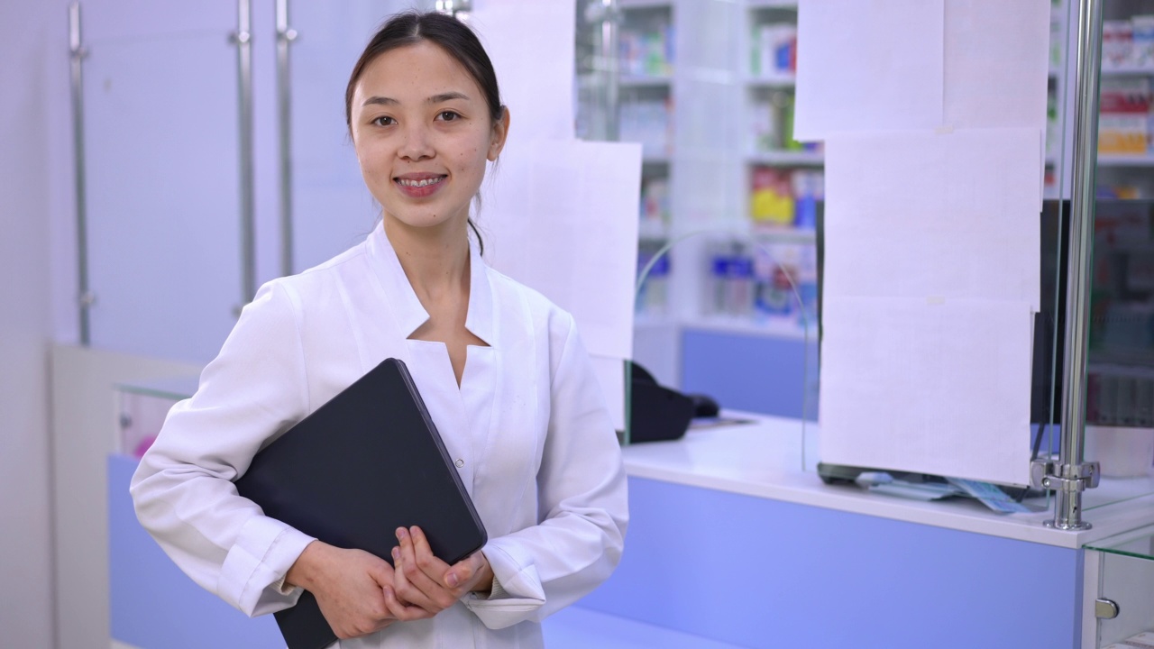 女亚洲药剂师收银员与数码平板在收银台在药店。积极自信的美丽年轻女子的肖像看着相机微笑站在药房室内。视频下载