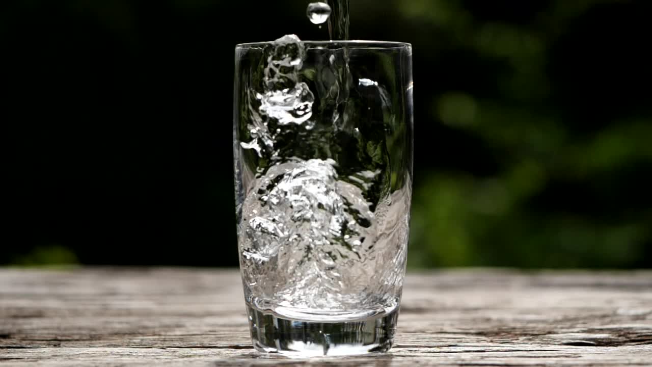 浇注的饮用水。特写镜头将净化的新鲜干净的饮用水倒入玻璃杯中，水在自然背景下飞溅，用慢动作柔和聚焦。饮酒、健康、自然的概念视频素材