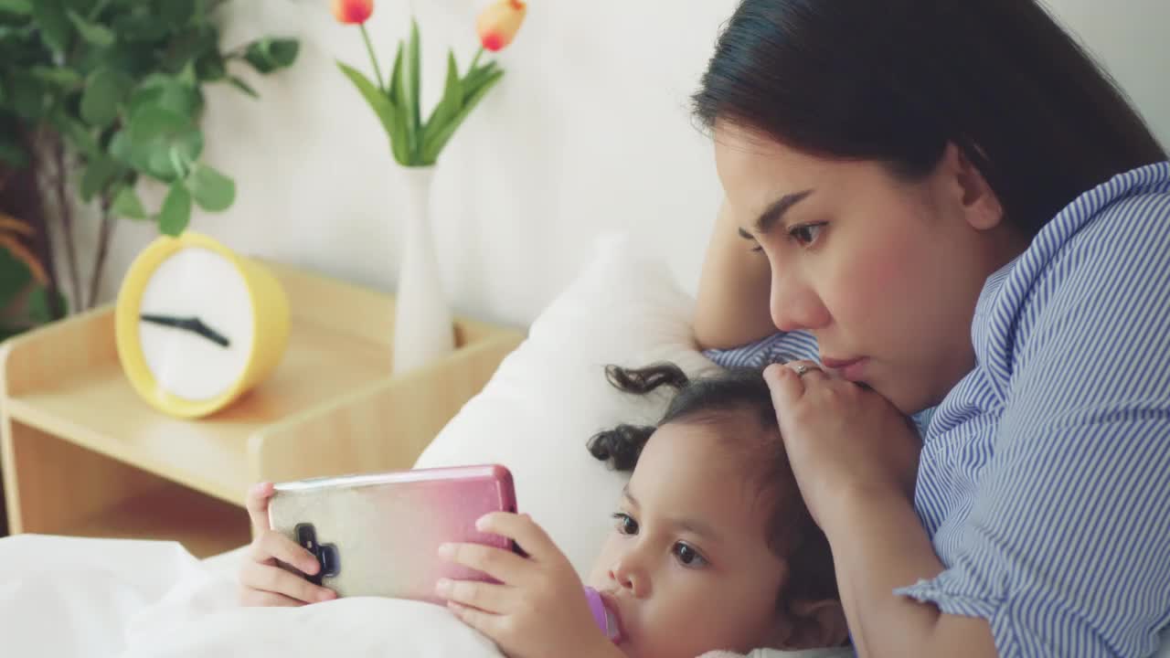 亚洲妈妈和宝宝躺在床上看智能手机视频素材