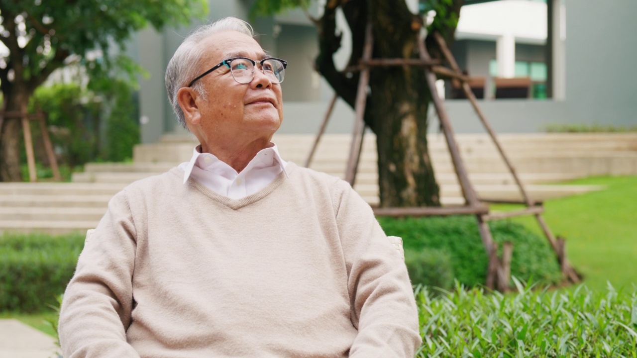 幸福生活亚洲退休老年老年男性坐在椅子上环顾四周感到平静和放松的绿色自然户外公园在一个阳光明媚的日子后练习散步。视频下载