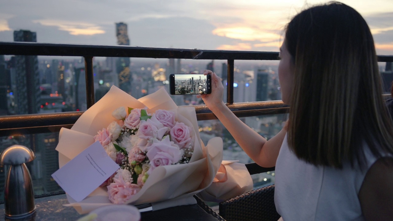年轻的亚洲女人拿着玫瑰花束庆祝生日在日落的屋顶在城市豪华的生活方式在户外晚餐微笑和快乐的晚餐浪漫的泰国曼谷视频下载
