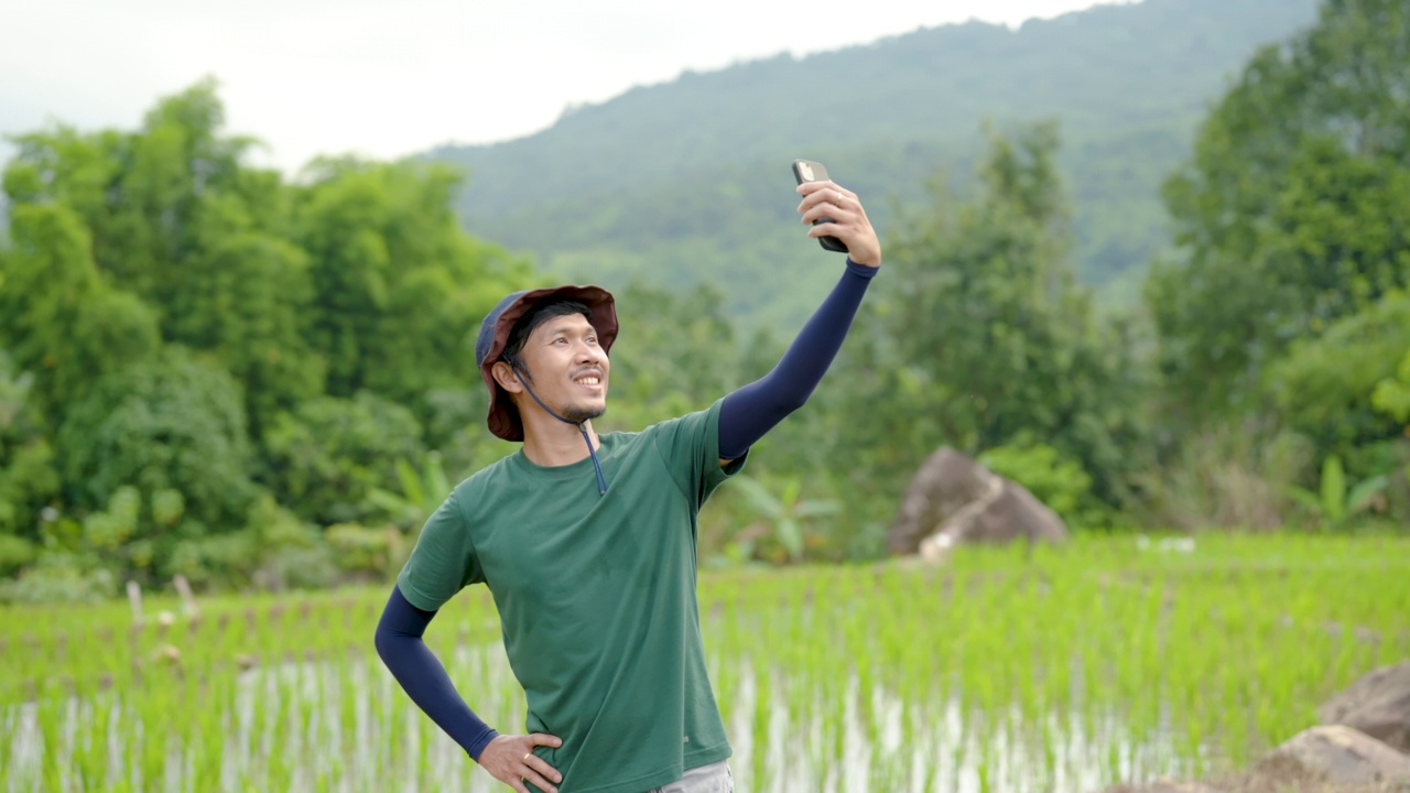 4K，留着小胡子的亚洲男人，高高的身材，戴着他最喜欢的太阳帽。他举起手机，与眼前的绿色田野自拍，开心地笑了。视频下载