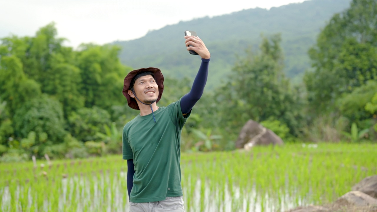 4K，留着胡子的亚洲人，高大的身材，戴着他最喜欢的太阳帽，他举起手机，和他面前的绿色田野自拍，开心地笑了。视频下载