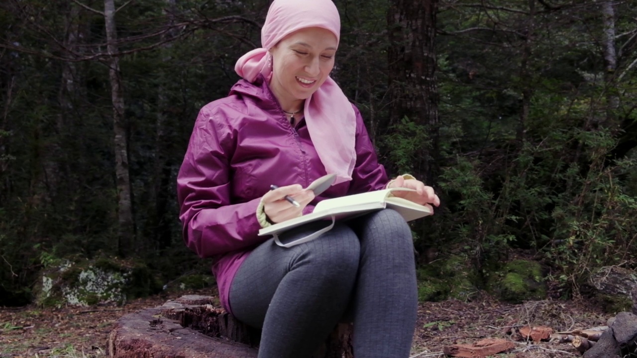 一名年轻的身患癌症、戴着粉色头巾的西班牙裔女性在树林里的篝火旁写日记。视频下载