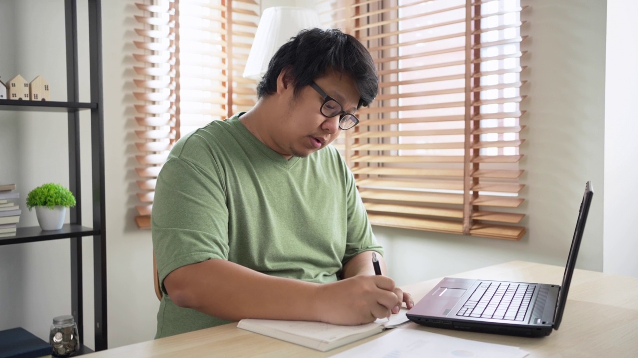 年轻的亚洲困倦男子在家里用笔记本电脑工作。努力在截止日期前完成工作的过度工作的自由职业者视频素材