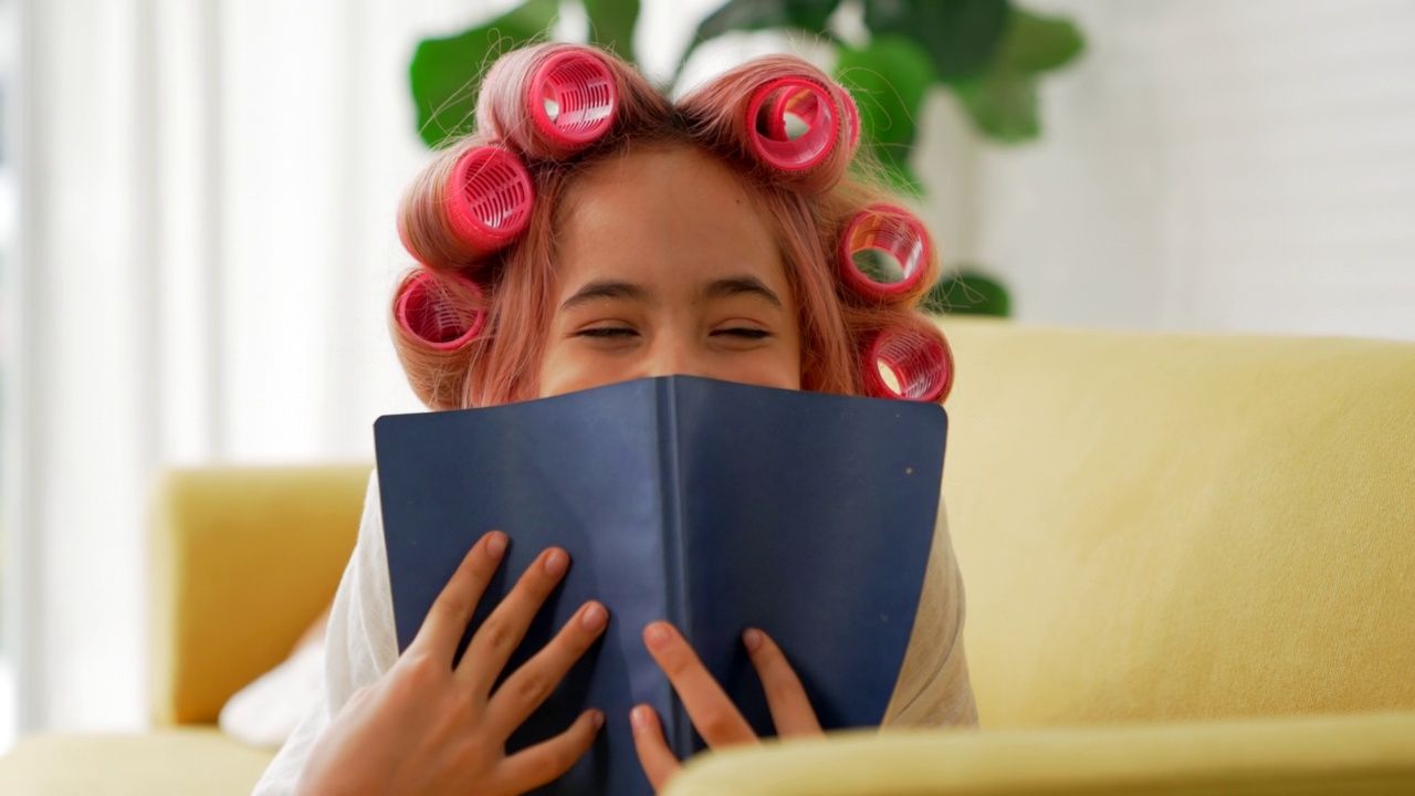 年轻开朗的女人带着卷发器，浓妆艳抹，手里拿着一本书，坐在家里的沙发上。化妆化妆在家。视频下载