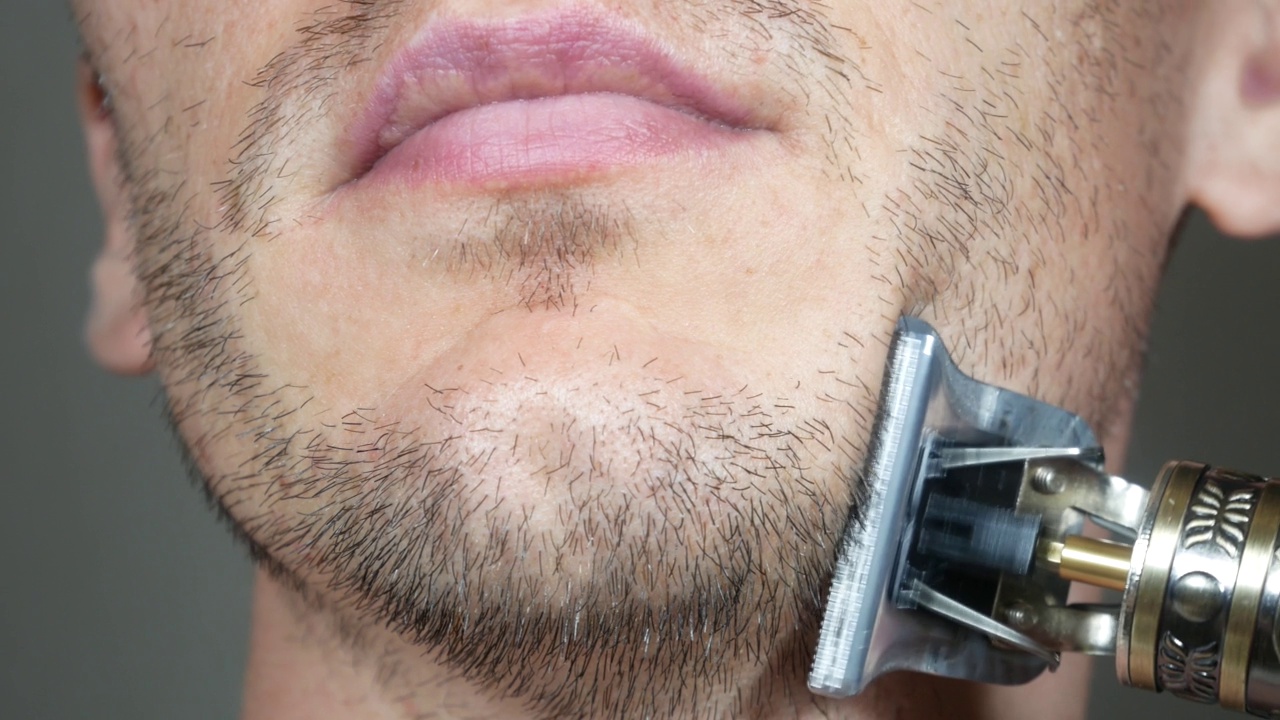 男子正在用电动金属剃须刀或干式剃须刀刮胡。男人脸上短而稀疏的胡须。头发生长的问题。胡子上有脱发区域的男子。胡子上的毛都没刮干净。视频下载
