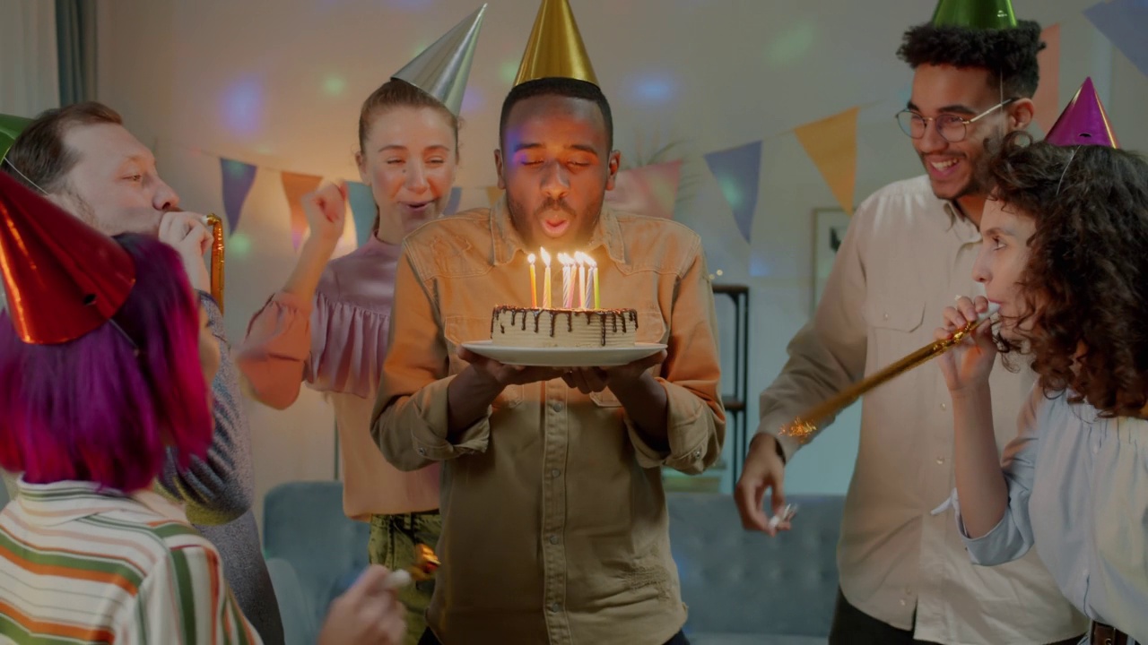 在家庭聚会上，当朋友们笑着鼓掌时，惊讶的非洲裔美国人吹着生日蛋糕上的蜡烛视频素材