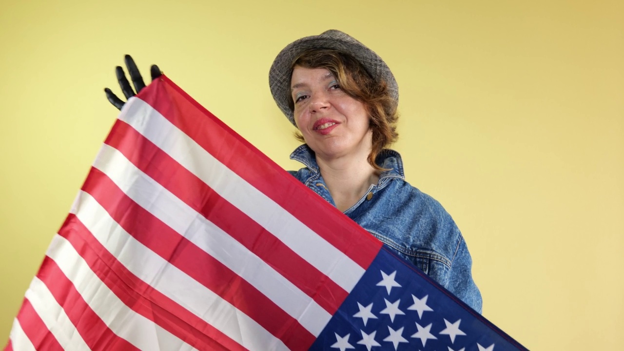 成年美国妇女在帽子和牛仔夹克挥舞美国国旗在孤立的黄色背景。女人的肖像视频下载