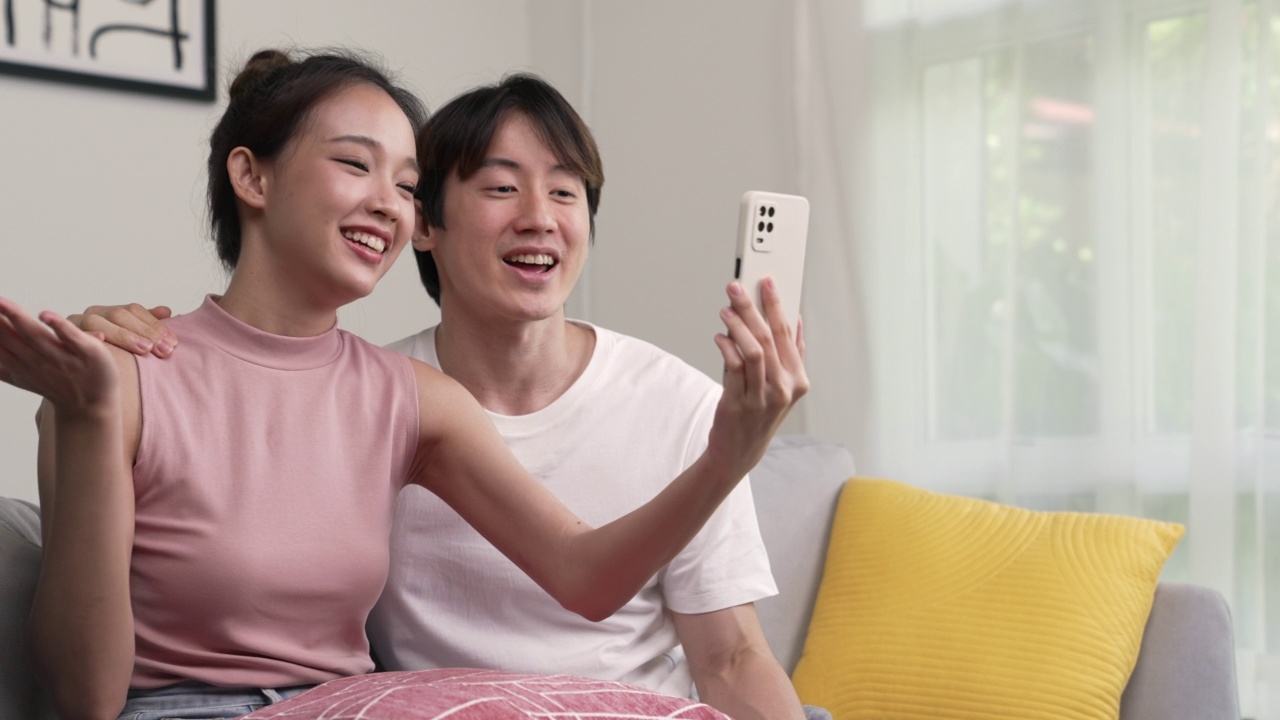 年轻的亚洲夫妇坐在一起工作，在家里用智能手机上网和朋友见面。视频素材