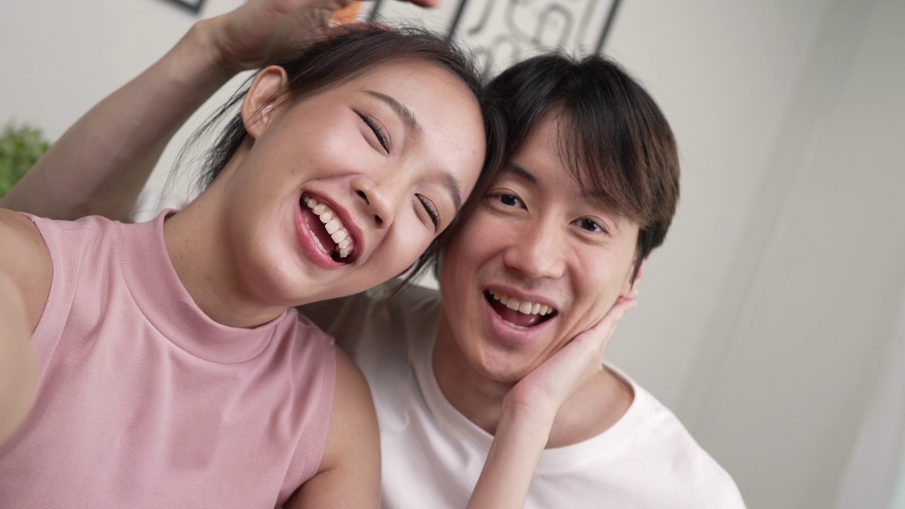 年轻的亚洲夫妇坐在一起看着镜头，在家里用智能手机与朋友和家人在线见面。视频素材