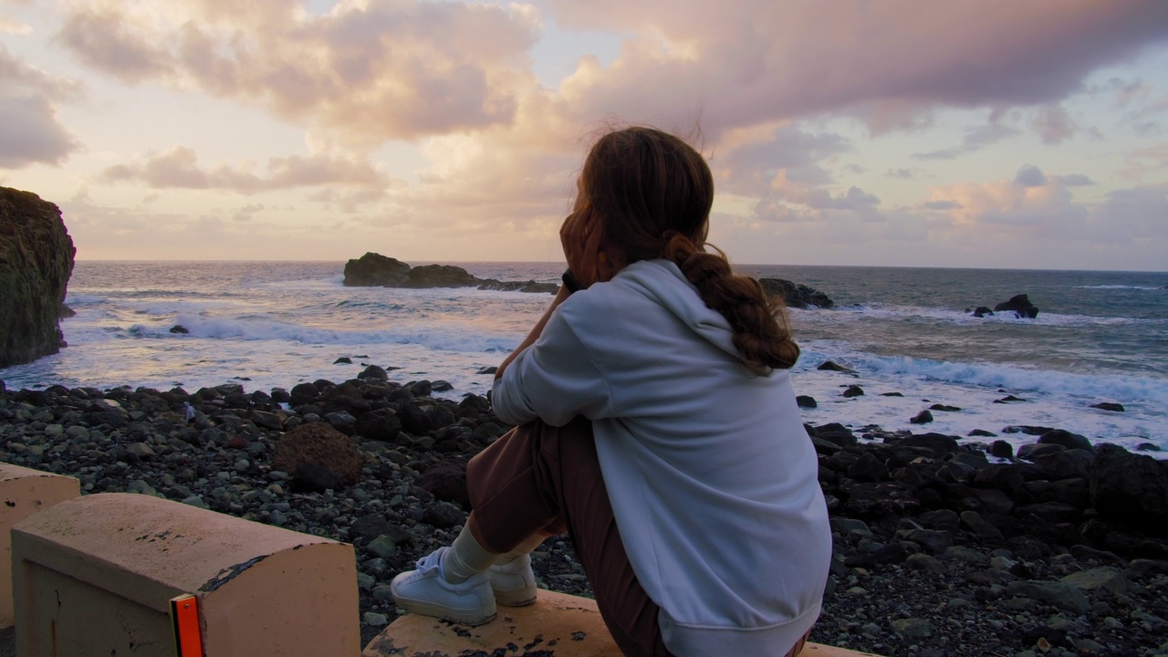 一个年轻女人的剪影坐在海边海滩附近的路边，看着日落在海洋，特内里费。后视图，女孩享受独自在火山岩石海岸，看日落视频素材