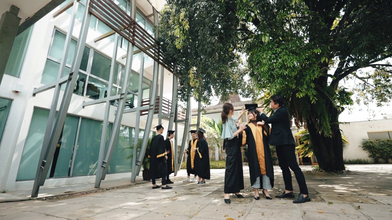 自豪的亚裔华裔父母在学校大楼外为穿着大学毕业服的女儿调整学位帽视频下载