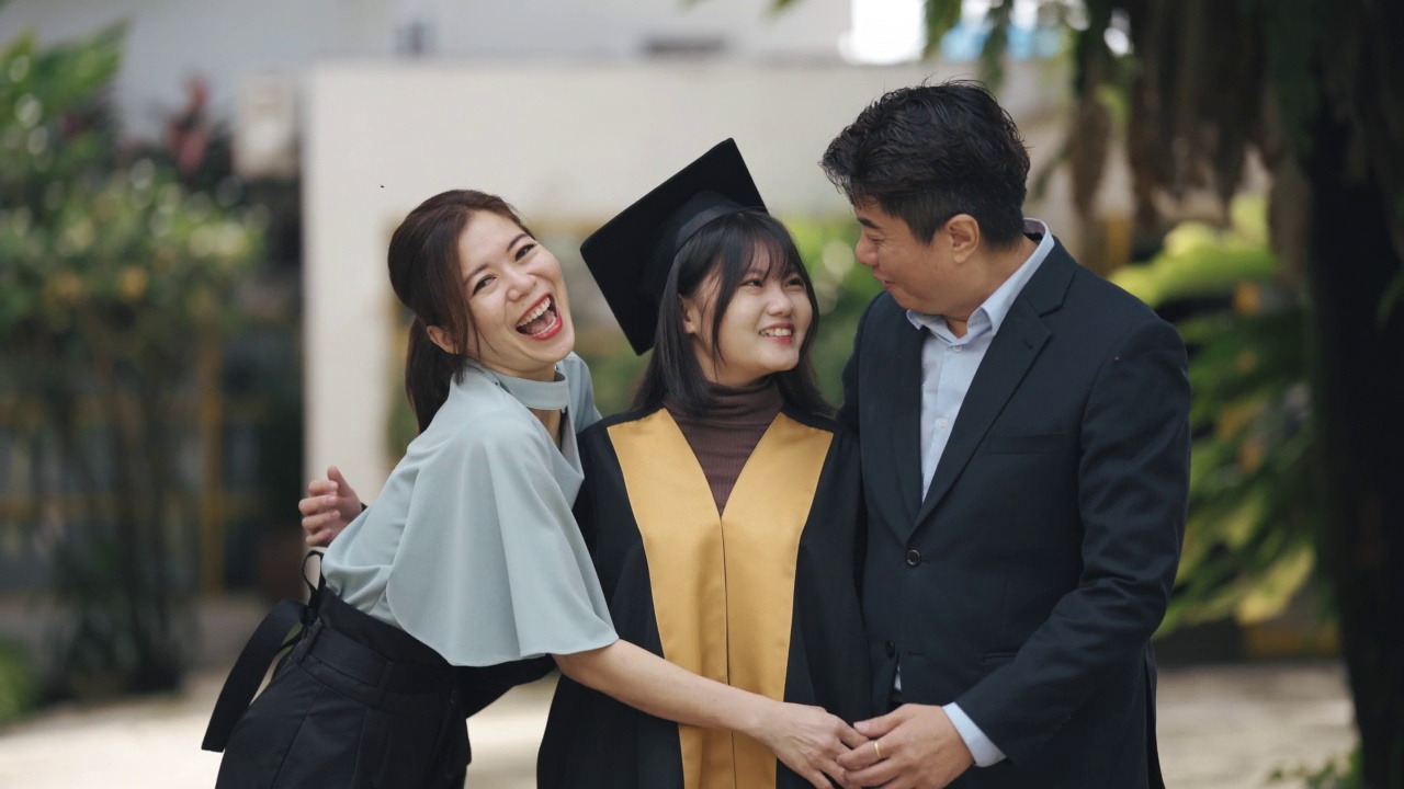 自豪的亚裔中国父母和他们的女儿穿着大学毕业礼服站在大学大楼外视频下载
