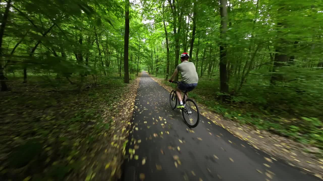 一名戴着防护头盔的男子正骑着自行车沿着森林中的柏油路前行。视频素材