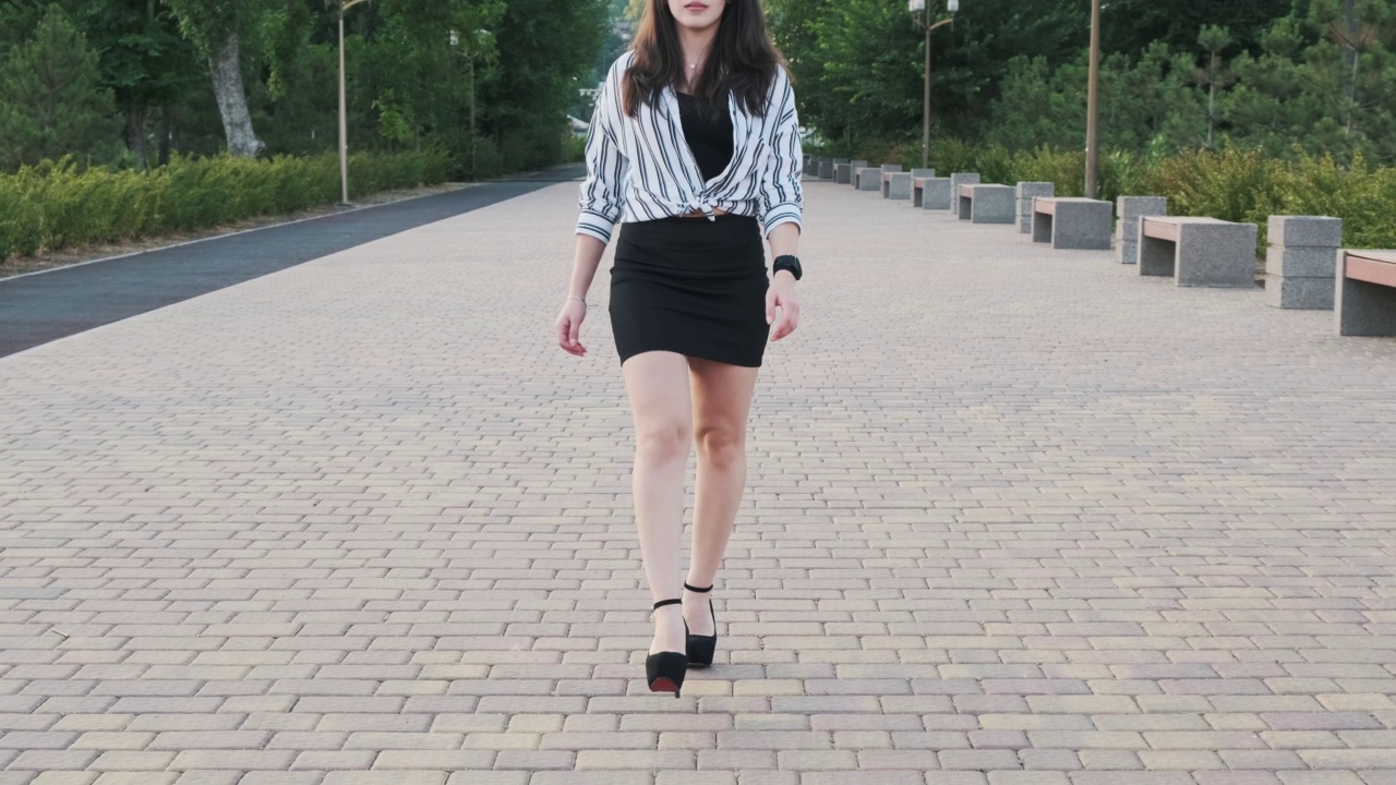 美丽的女孩穿着黑色的高跟鞋，黑色的裙子和条纹衬衫走在公园里。前视图。慢动作视频素材